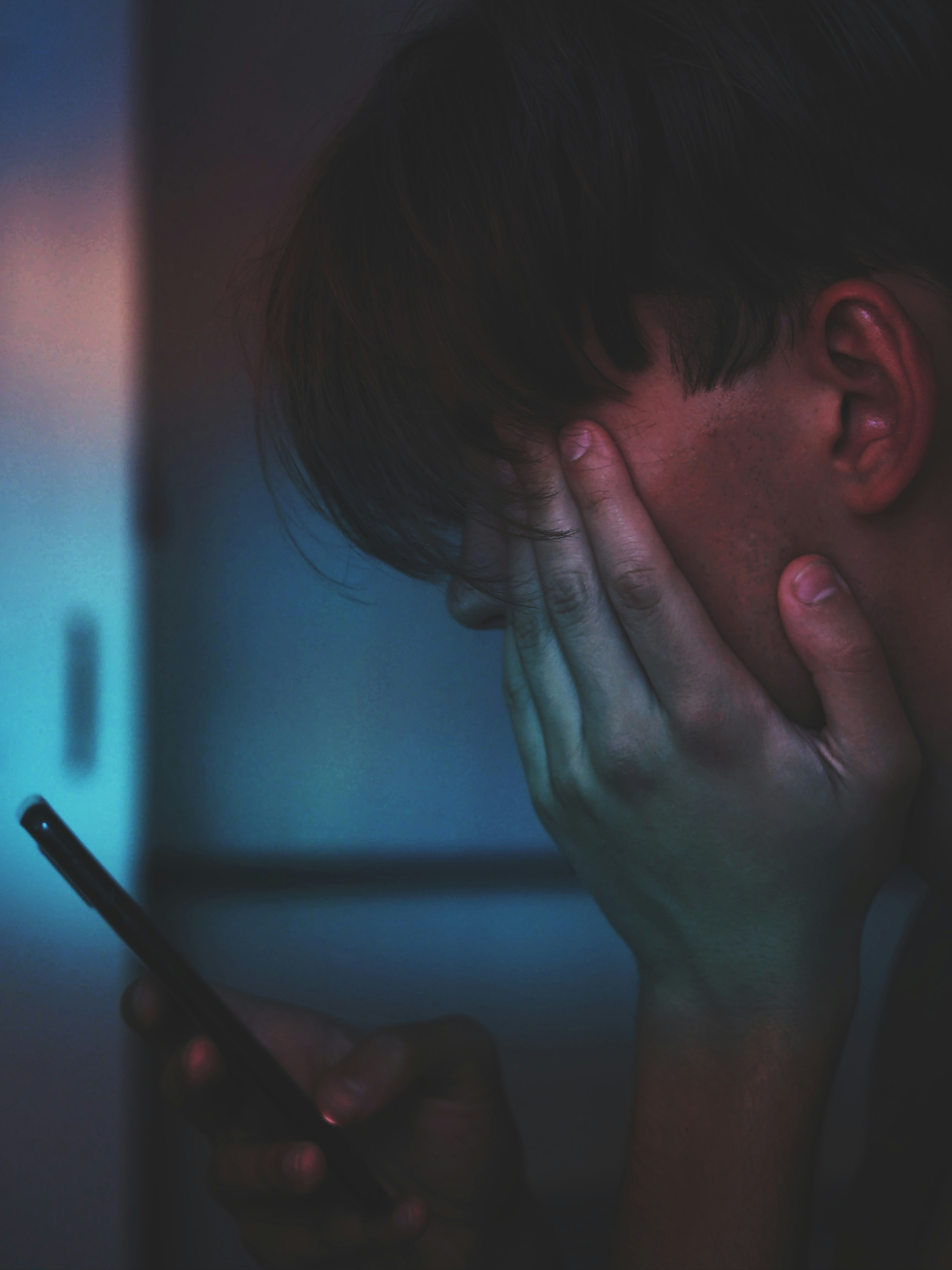 Adolescente angustiado, con la mano sobre la cara, mirando el celular en la penumbra