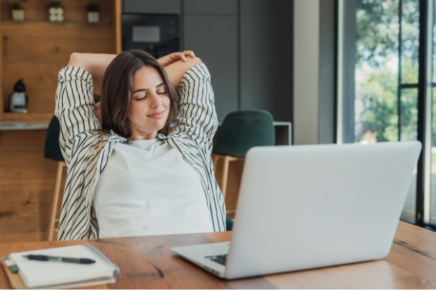 Una mujer sentada frente a una notebook se estira con las manos detrás de la cabeza en un ambiente con ventanal