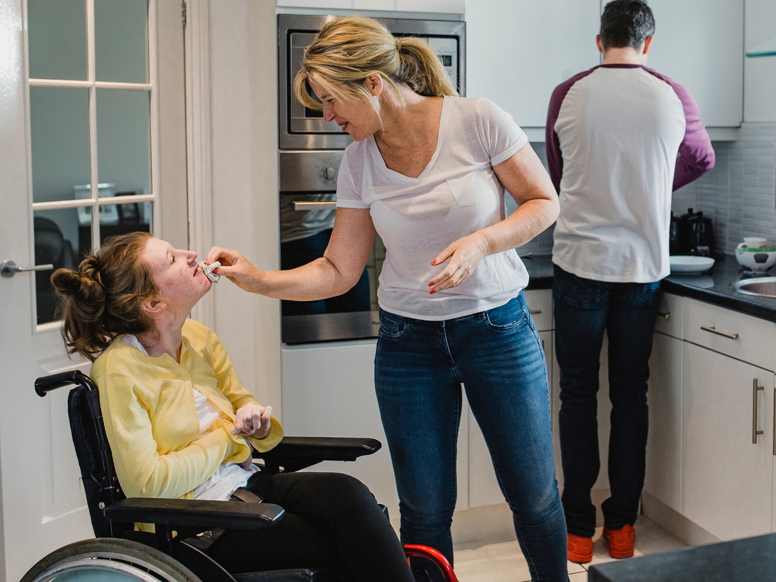 Una madre le acerca la mano a la cara a su hijo, sentado en silla de ruedas, en una cocina asistiendolo