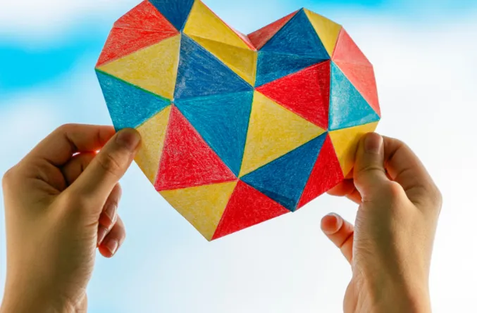 Manos sosteniendo un corazón de papel formado por triángulos de colores contra el cielo