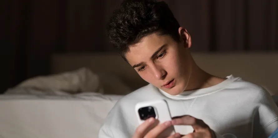 Adolescente sentado en la cama, concentrado en la pantalla de su celular