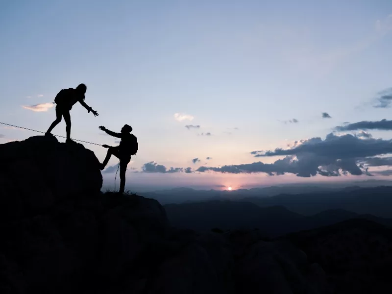 Silueta de una persona ayudando a otra a subir a la cima de una montaña al atardecer