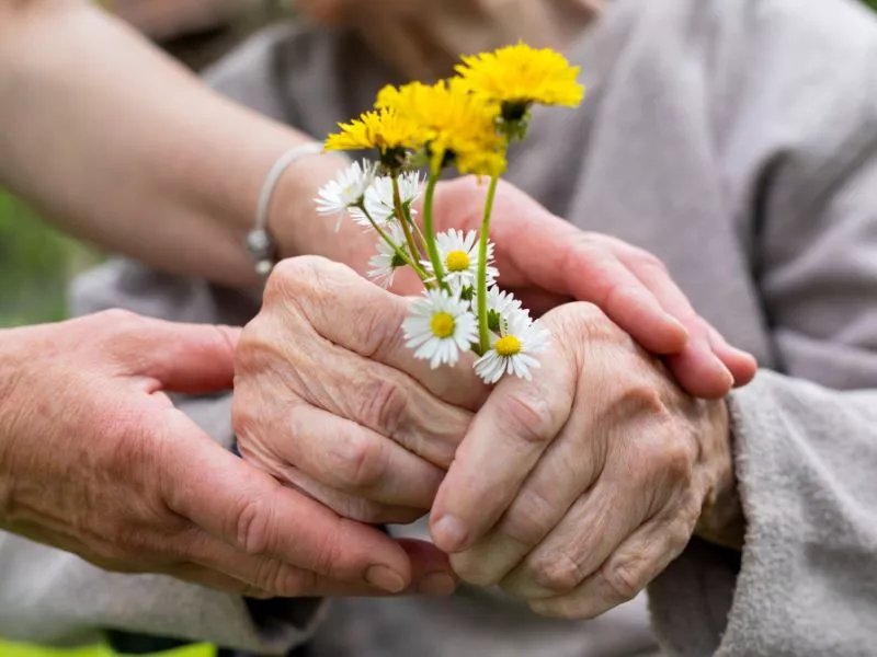 Manos jóvenes sosteniendo las manos de una persona mayor, que tiene un ramito de flores amarillas