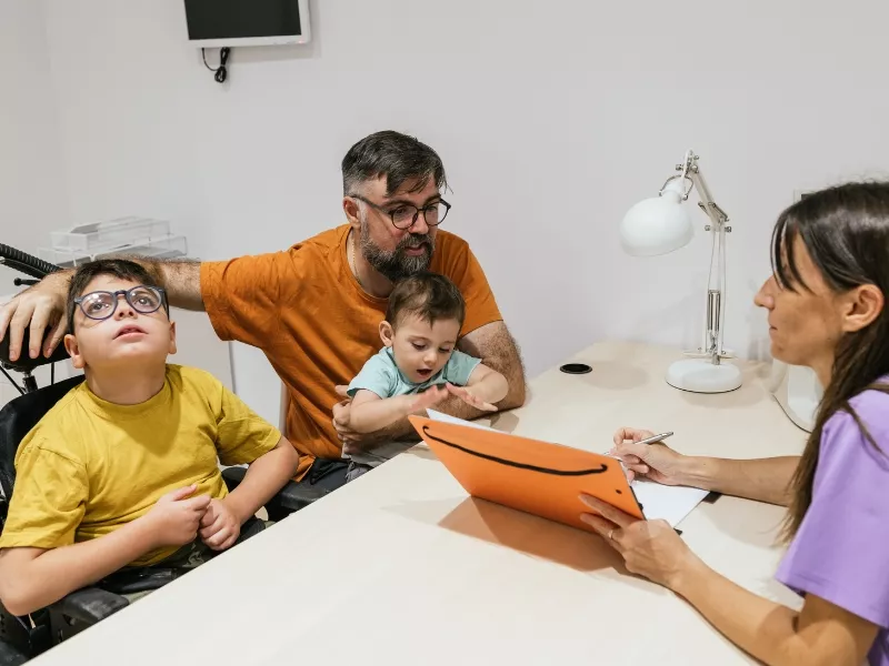 Hombre con dos niños, uno en silla de ruedas, conversando con una profesional en un consultorio