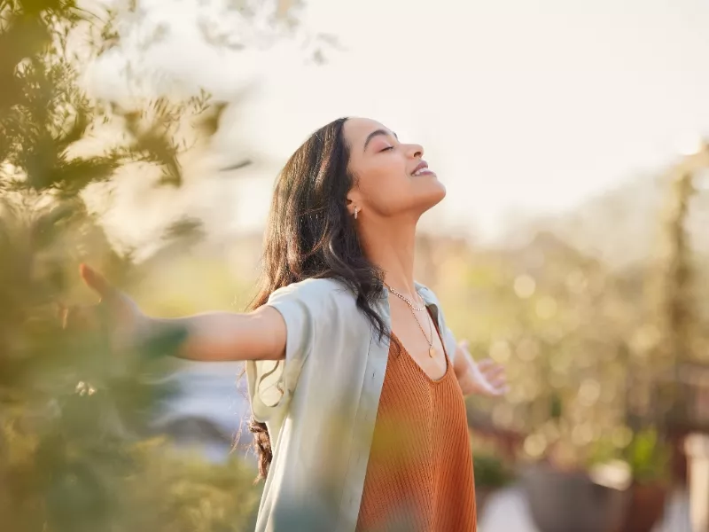 Mujer con los brazos abiertos y los ojos cerrados, respirando al aire libre entre árboles