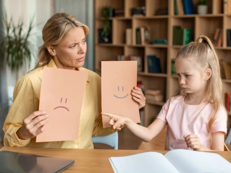 Mujer mostrando a una niña tarjetas con caras triste y contenta para que elija una emoción