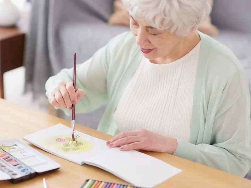 Mujer mayor pintando con acuarelas en una mesa, concentrada en su dibujo