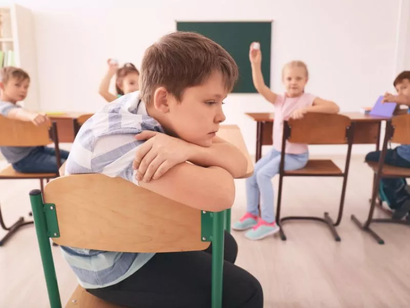 Niño triste y aislado en su pupitre mientras sus compañeros participan en clase