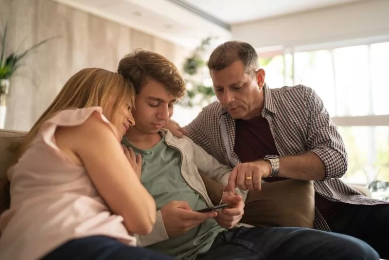 Dos adultos acompañan a un adolescente en el sillón mientras él mira el celular