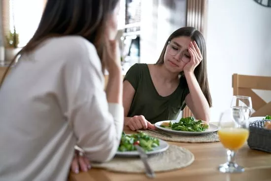 Adolescente desganada frente a su plato en la mesa familiar, mientras una mujer la mira
