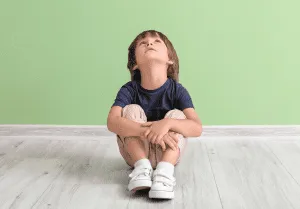 Niño sentado en el piso abrazando sus rodillas, mirando hacia arriba, frente a una pared verde