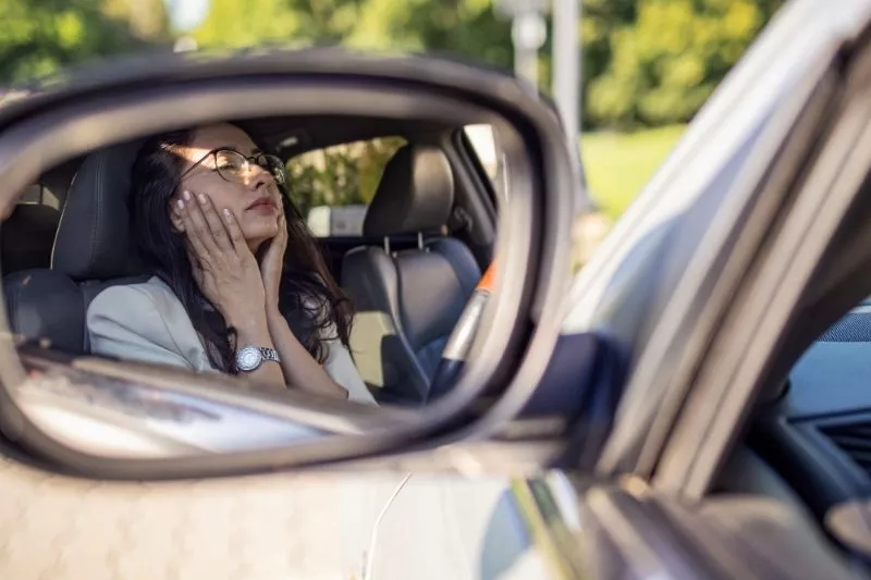 Mujer agobiada dentro de un auto, con las manos en la cara, reflejada en el espejo retrovisor